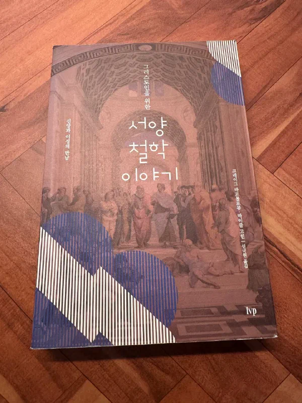 『그리스도인을 위한 서양 철학 이야기』 표지. 라파엘로의 &lsquo;아테네 학당&rsquo;을 붉은 톤으로 깔고 파란 스트라이프 도형을 얹은 IVP판 표지가 나무 바닥 위에 놓여 있다