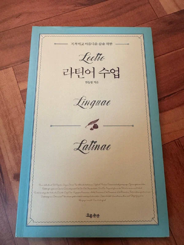 나무 바닥에 놓인 『라틴어 수업』의 청록색 표지. 가운데에 「지적이고 아름다운 삶을 위한」이라는 문구와 필기체로 쓴 「Lectio」, 「Linguae」, 「Latinae」가 배치되어 있고, 그 사이에 한글 제목 「라틴어 수업」과 「한동일 지음」이 적혀 있다. 아래쪽에는 깃털 펜과 잉크병 그림, 그리고 출판사 흐름출판이 있다