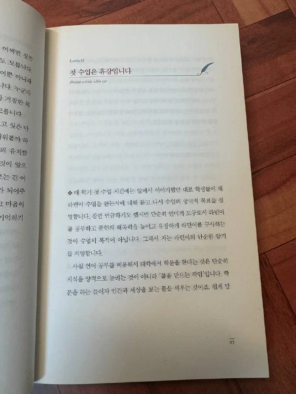 책 27쪽. 「Lectio II」 아래에 「첫 수업은 휴강입니다」라는 제목과 라틴어 「Prima schola alba est」가 적혀 있고, 오른쪽에 깃털 펜 장식이 있다. 본문에는 매 학기 첫 수업에 학생들이 왜 라틴어 수업을 듣는지 듣고 나서 수업의 궁극적 목표를 설명한다는 이야기, 그리고 「대학에서 학문을 한다는 것은 지식을 양적으로 늘리는 것이 아니라 틀을 만드는 작업」이라는 문장이 이어진다