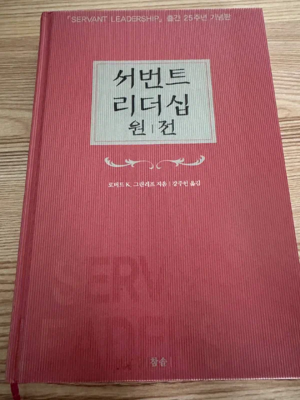 붉은 세로줄 무늬 양장 표지에 &lsquo;서번트 리더십 원전&rsquo;이라는 제목이 있고 아래에 로버트 K. 그린리프 지음, 강주헌 옮김, 출판사 참솔이 적힌 책이 나무 책상 위에 놓여 있다