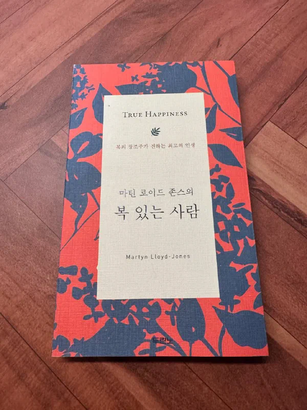 붉은색과 남색 꽃무늬 표지에 &lsquo;TRUE HAPPINESS&rsquo;, &lsquo;마틴 로이드 존스의 복 있는 사람&rsquo;이라고 적힌 두란노 책이 나무 바닥 위에 놓여 있다
